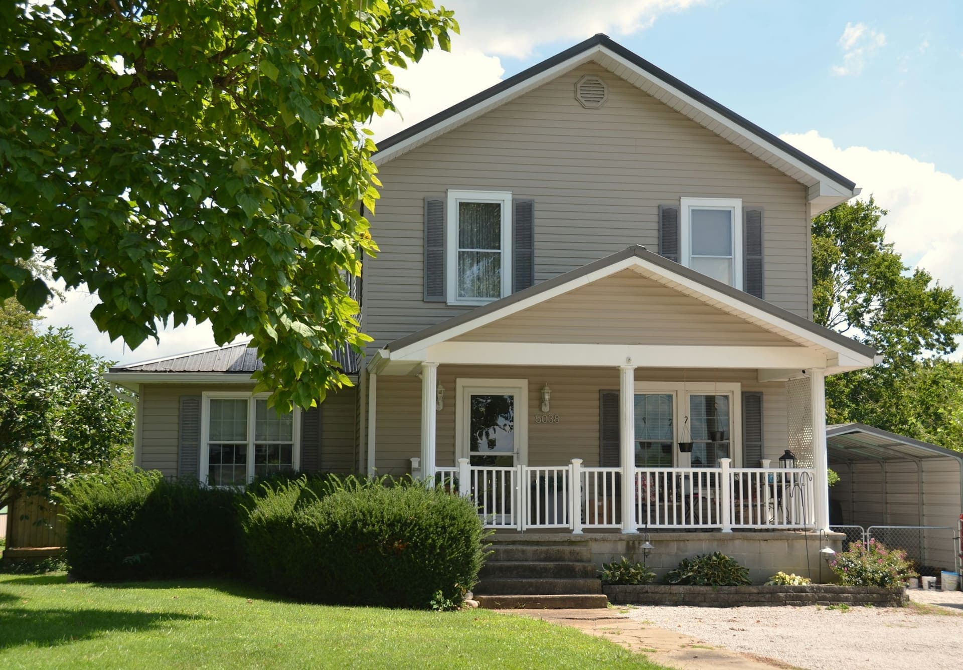 Two-story house with beige siding and a white front porch beside a large maple tree
